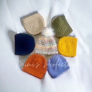 Könnte beinhalten: Eine Sammlung handgefertigter Häkelmützen in verschiedenen Farben, darunter Beige, Marineblau, Orange, Gelb, Olivgrün und Hellblau. Eine Mütze hat ein mehrfarbiges Design und einen weißen Bommel. Der Text "Mimis Perfection" ist sichtbar.