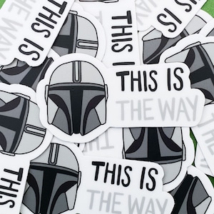 May include: A set of stickers featuring a Mandalorian helmet and the phrase "This is the Way". The stickers are white with black text and a grey helmet design.