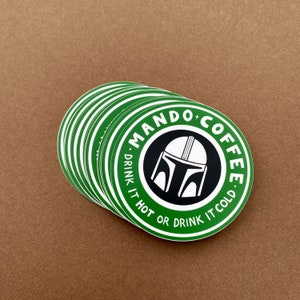 May include: A pile of green and white circular stickers featuring the words "MANDO COFFEE" and a Mandalorian helmet design. The text "DRINK IT HOT OR DRINK IT COLD" is also printed on the stickers. The stickers are on a brown surface.