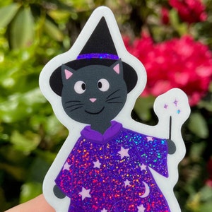 May include: A black cat wearing a purple and white sparkly wizard costume with a black witch hat. The cat is holding a wand with stars coming off of it.
