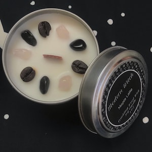 May include: A silver tin with a black label that reads "Modern Witch Maple Latte Soy Candle" with a design of small black squares. The lid is open and reveals a white candle with coffee beans, crystals, and a cinnamon stick.