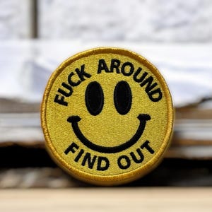 May include: Yellow embroidered patch with a smiling face design. The patch features the text "FUCK AROUND" above the eyes and "FIND OUT" below the mouth in black capital letters. The patch is round and has a yellow border.