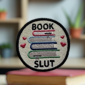 May include: Embroidered patch with the text "BOOK SLUT" and a stack of books. The patch is round with a black border and features red hearts. The books are in shades of red, blue, and green.