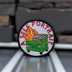 May include: Embroidered patch with the text "A SELF PORTRAIT" around the edge. The patch features a green dumpster with a smiling face and flames. The dumpster is filled with gray trash. The patch has a black border.