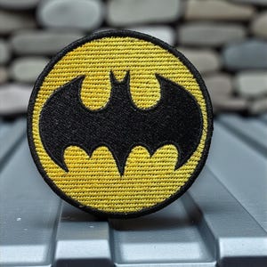 May include: A round, embroidered patch featuring the iconic Batman logo. The patch has a black bat symbol on a yellow background with a black border. The patch is placed on a corrugated metal surface.