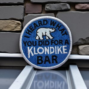 May include: A round, blue embroidered patch with white text and a polar bear graphic. The text reads "I HEARD WHAT YOU DID FOR A KLONDIKE BAR". The patch is set against a textured stone wall.