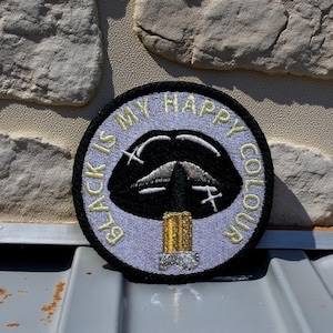 May include: Round embroidered patch with the text "BLACK IS MY HAPPY COLOUR" in gold thread. The patch features a black lip design with white accents and a gold lipstick. The patch is on a light purple background.