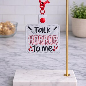 May include: A white keychain with the text "Talk Horror To Me" in red and black, featuring knife illustrations. The keychain has a red clasp and button, displayed on a gold stand.
