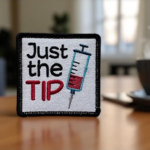 May include: Embroidered patch with the text "Just the TIP" in black and red, featuring a syringe graphic. The patch is square with a black border. A cup of coffee sits in the background.