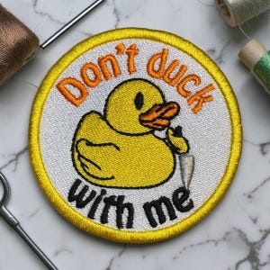 May include: Embroidered patch featuring a yellow cartoon duck holding a knife. The patch has a white background with the text "Don't duck with me" in orange and black. The patch is round with a yellow border.