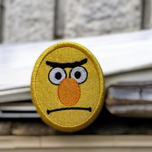 May include: Embroidered yellow patch featuring a grumpy face with black eyebrows, white eyes, and an orange nose. The patch has a black stitched mouth and is likely an iron-on or sew-on accessory for clothing or bags.