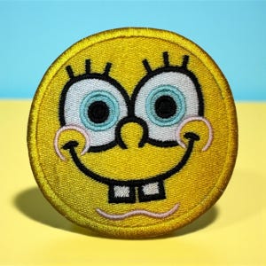 May include: A yellow embroidered patch featuring the face of a cartoon character with large blue eyes, black eyelashes, and a wide smile with two square teeth. The patch has a circular shape and a stitched border.