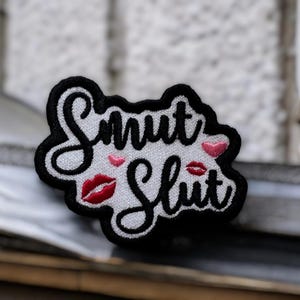 May include: Embroidered patch with the words "Smut Slut" in black script, outlined in black, on a white background. The patch features red lips and pink heart accents. The patch is a decorative item.