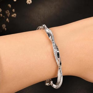 Op de afbeelding: Een zilverkleurige armband met een gedraaid, spiraalvormig ontwerp. Het oppervlak van de metalen band wisselt af tussen gladde, gepolijste delen en getextureerde, sprankelende segmenten die het licht vangen, wat een continu touwachtig effect creëert.