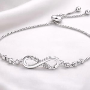 Könnte beinhalten: Ein silberfarbenes Armband mit einem Unendlichkeitssymbol in der Mitte, verziert mit kleinen, funkelnden Steinen. Das verstellbare Kettenarmband hat ein zartes Design und ist vor einem weißen, seidigen Hintergrund platziert.
