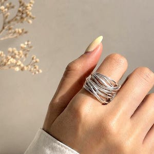 Könnte beinhalten: Ein silberner Ring mit einem gewebten Design. Der Ring hat mehrere Metallstränge, die sich verflechten, um ein strukturiertes, geschichtetes Aussehen zu erzeugen. Der Ring wird an einer Hand getragen, mit einem neutralen Hintergrund.