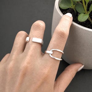 May include: Three silver rings are shown on a hand. One ring features a rectangular design, and another has a double-banded design. The rings have a simple, modern style with a polished finish. The rings are likely made of sterling silver.