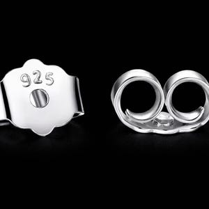 May include: Two silver earring backs, one with a post hole and the number "925" stamped on it. The other earring back has two loops. Both are against a black background.