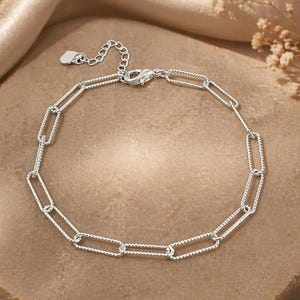 May include: A silver link bracelet with a lobster clasp. The bracelet has rectangular links with a textured, rope-like pattern. It is displayed on a textured, light brown surface. The bracelet's design is elegant.