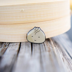 May include: A small, beige, smiling dumpling pin sits on top of a wooden surface. The pin is in front of a stack of bamboo steamer baskets.
