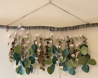 May include: A decorative wall hanging featuring a natural branch with hanging eucalyptus leaves, pink flowers, and a small butterfly accent. The arrangement is suspended by twine, creating a rustic and organic aesthetic. The leaves are various shades of green and brown.