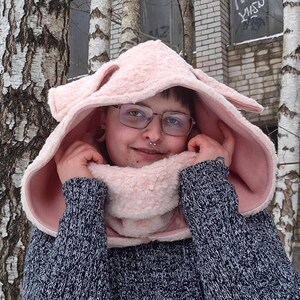 Könnte beinhalten: Ein hellrosa Kapuzenschal mit Hasenohren, getragen über einem dunkelblauen Strickpullover. Der Schal ist aus einem weichen, flauschigen Material gefertigt. Die Innenseite der Kapuze und des Schals ist dunkler rosa.