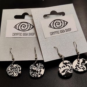 Könnte beinhalten: Zwei Paar schwarze und weiße Ohrringe mit Wirbelmuster an silbernen Haken. Die Ohrringe werden auf einer weißen Karte mit dem aufgedruckten Text "Cryptic Idea Shop" präsentiert.