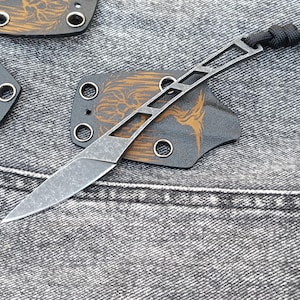 Peut inclure: Un petit couteau de poche noir avec un design unique à cadre ouvert. Le couteau a une poignée noire avec un motif gravé brun et un trou pour un cordon. Le couteau est posé sur une surface en jean gris.