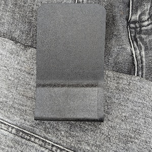 Op de afbeelding: Een rechthoekige, zwarte telefoonhoes met een getextureerd oppervlak en afgeronde hoeken. De hoes wordt getoond tegen een grijze denim achtergrond, wat de donkere kleur en het tastbare oppervlak benadrukt. De hoes heeft een kleine lip aan de onderkant.
