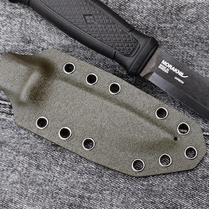 May include: A green leather knife sheath with metal eyelets. The sheath is lying on a grey denim fabric.