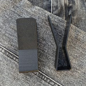 Puede incluir: Dos objetos negros sobre un fondo de mezclilla gris. Uno es un bloque rectangular con una superficie texturizada, dividido en dos tonos de negro. El otro es un objeto negro abstracto en forma de V.