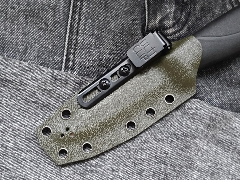 May include: A green knife sheath with a black clip and multiple holes for attaching to a belt. The sheath is made of a textured material and has a smooth finish.