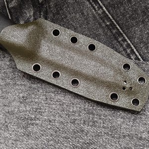 May include: A green leather knife sheath with multiple metal eyelets. The sheath is lying on a dark gray denim fabric.