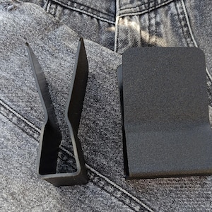 May include: Two black metal objects rest on a denim surface. One is a money clip with a textured finish, and the other is a stylized, open-ended shape. The objects are likely accessories or tools.