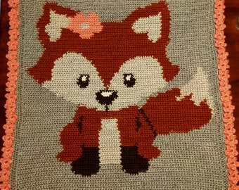 May include: A crocheted fox blanket with a gray background and a pink border. The fox is wearing a red scarf and has a pink flower on its head.