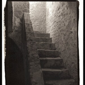 May include: A black and white photograph of a stone staircase leading up to a doorway. The staircase is made of rough-hewn stone and the walls are made of a similar material. The photo is taken from a low angle, looking up at the staircase.