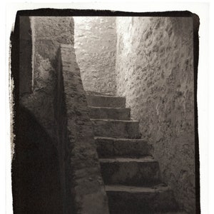 May include: A black and white photograph of a stone staircase leading up into a dimly lit room. The staircase is made of rough-hewn stone and has a handrail on the left side. The room is lit by a single window at the top of the stairs.