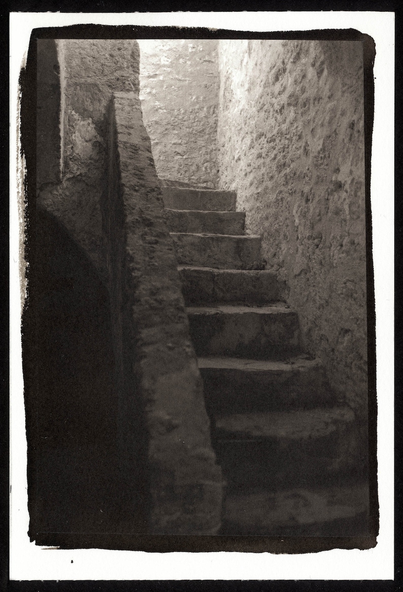 May include: A black and white photograph of a stone staircase leading up to a doorway. The staircase is made of rough-hewn stone and the walls are made of a similar material. The photo is taken from a low angle, looking up at the staircase.