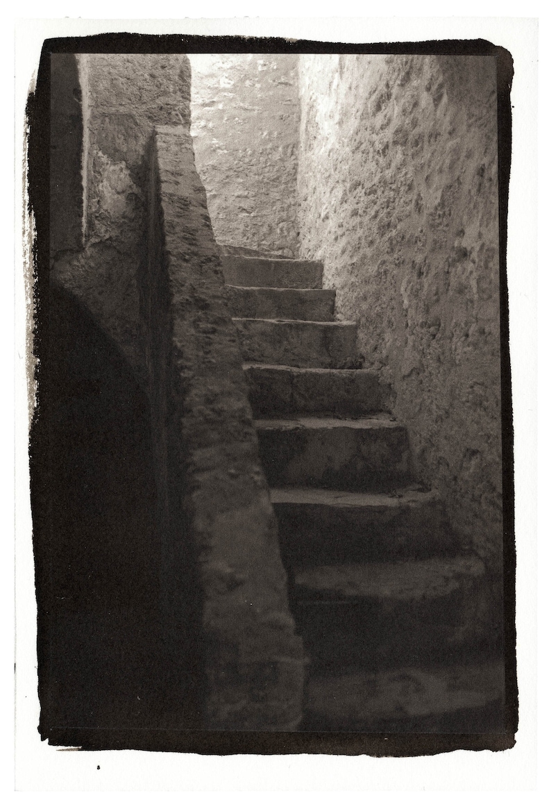 May include: A black and white photograph of a stone staircase leading up into a dimly lit room. The staircase is made of rough-hewn stone and has a handrail on the left side. The room is lit by a single window at the top of the stairs.