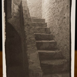 May include: A black and white photograph of a stone staircase leading up to a doorway. The staircase is in a narrow, enclosed space with rough stone walls.