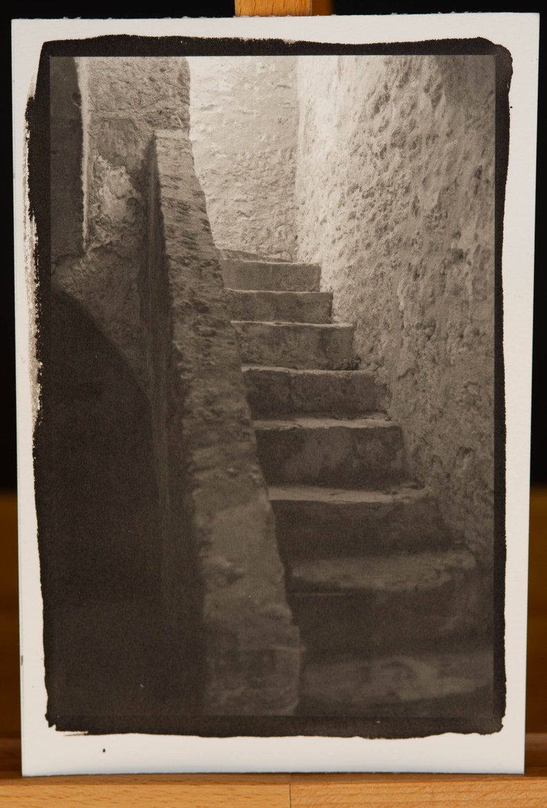 May include: A black and white photograph of a stone staircase leading up to a doorway. The staircase is in a narrow, enclosed space with rough stone walls.