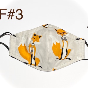 May include: A beige face mask with black adjustable straps. The mask features a repeating pattern of orange foxes wearing glasses and bowties. The text "F#3" is printed in brown at the top of the image.