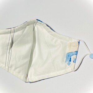 May include: White face mask with blue and white abstract print. The mask has adjustable ear loops and a nose wire.