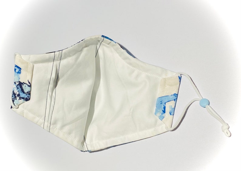 May include: White face mask with blue and white abstract print. The mask has adjustable ear loops and a nose wire.