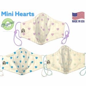 May include: Three white face masks with different colored heart patterns. The top mask has light purple hearts, the middle mask has light blue hearts, and the bottom mask has light blue hearts. All masks have a small brown logo on the left side and are made in the USA.