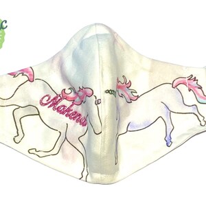 May include: White face mask with two unicorns, one with the name "Makenna" written on it. The mask has white elastic straps with pink bead accents. The mask is made in the USA.