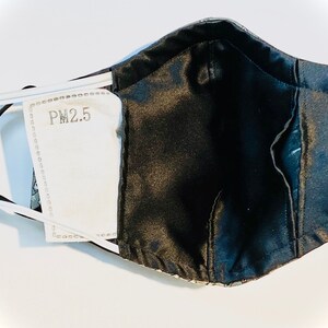 Peut inclure: Un masque facial noir avec une doublure &agrave; motifs blancs et gris. Le masque a une poche pour un filtre PM2.5, visible sur l'image.