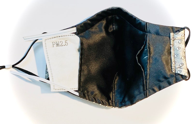 Peut inclure: Un masque facial noir avec une doublure &agrave; motifs blancs et gris. Le masque a une poche pour un filtre PM2.5, visible sur l'image.