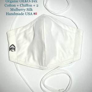 May include: A white face mask with adjustable white straps. The mask is made of multiple layers of fabric, including cotton, chiffon, and mulberry silk. The text on the image reads "5-Layer 1000 T.C Organic OEKO-Tex Cotton + Chiffon + 2 Mulberry Silk Handmade USA".