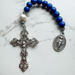 Puede incluir: Un rosario de madera azul con un crucifijo de plata y una medalla de plata de la Virgen María. El rosario tiene una perla blanca.